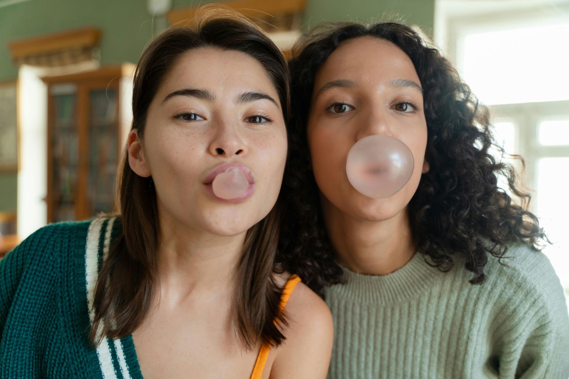 Two people blowing bubble gum bubbles in front of a window. One in a green sweater, one in a orange top.