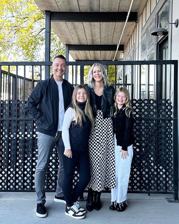 Family poses in front of a metal fence. Father wears a jacket. Mother in a skirt and jacket. Two children.