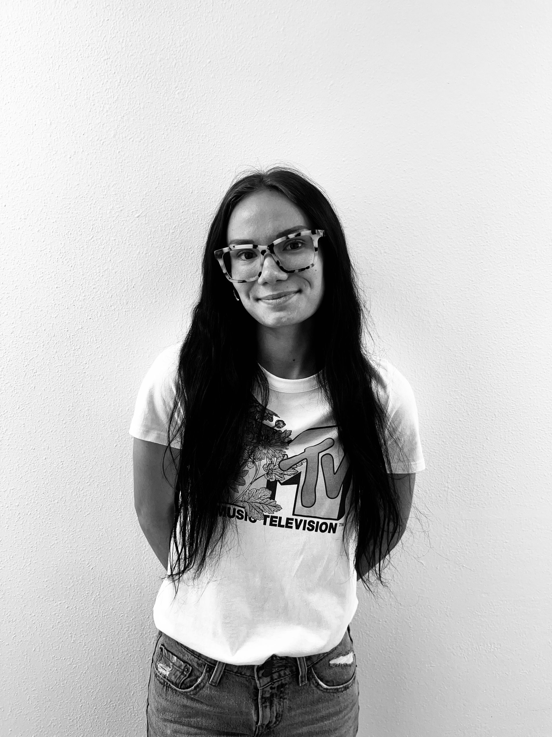 Person with long hair and glasses standing against a plain wall, wearing a white graphic T-shirt and smiling.