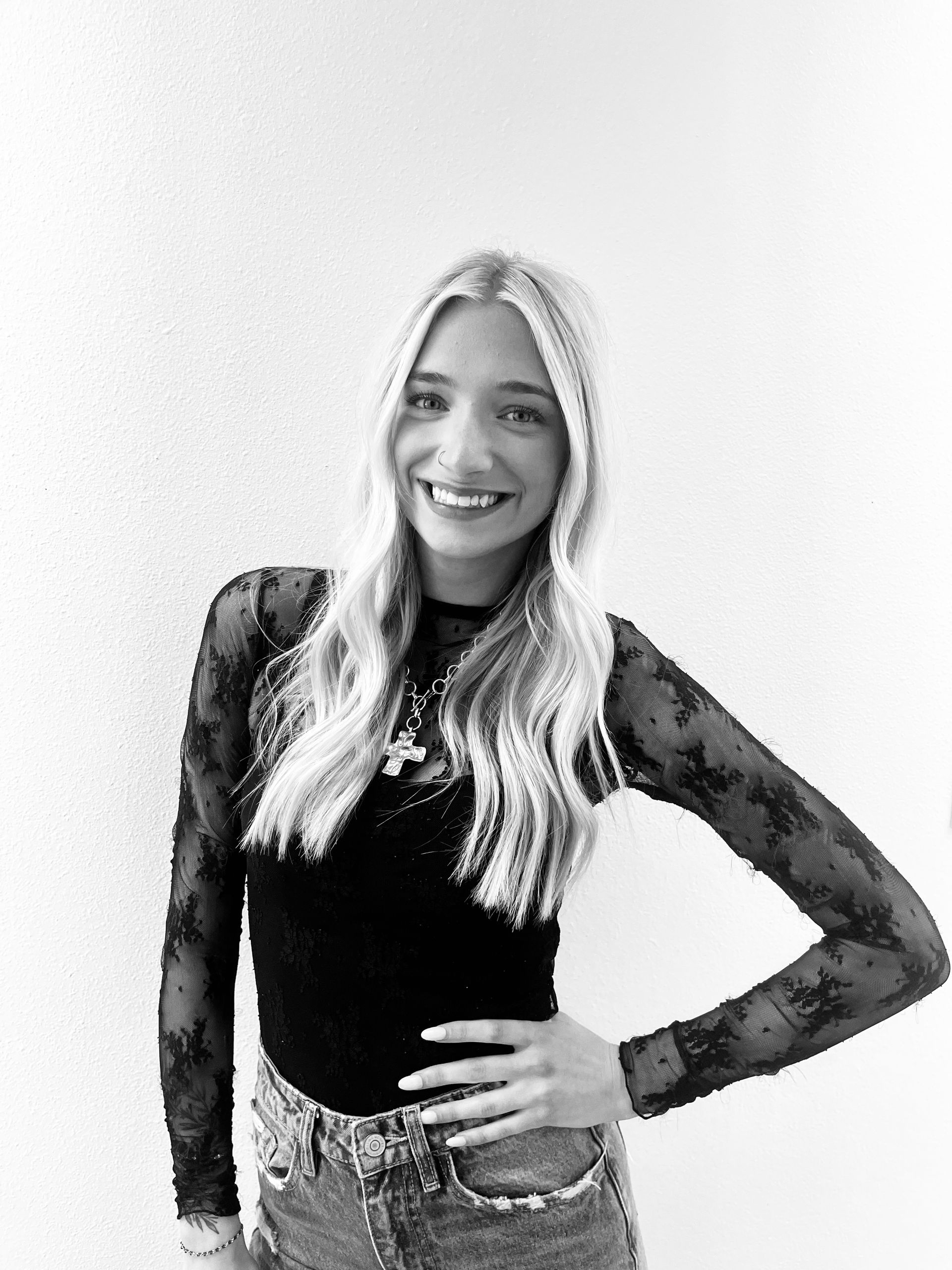 Woman with long dark hair, nose ring, and cross earring, smiling, in front of a white wall.