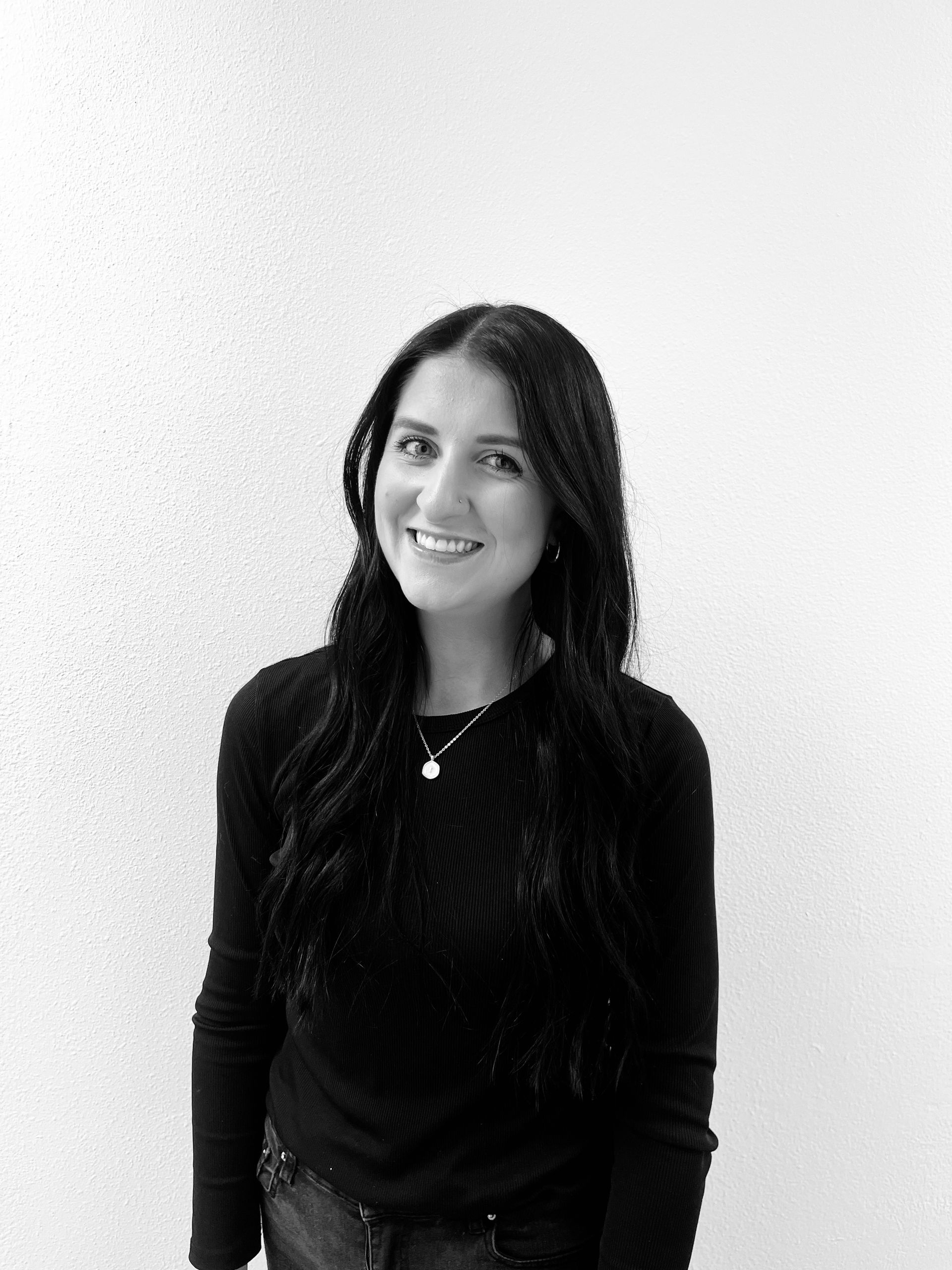 Smiling woman in a black top standing against a light wall.
