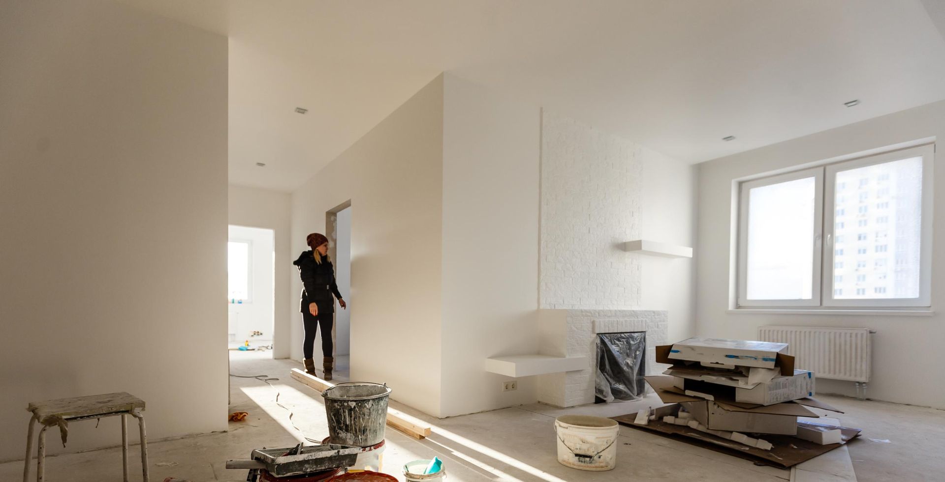 Person in black clothing stands in a bright, white-painted room under renovation. Construction materials litter the floor.