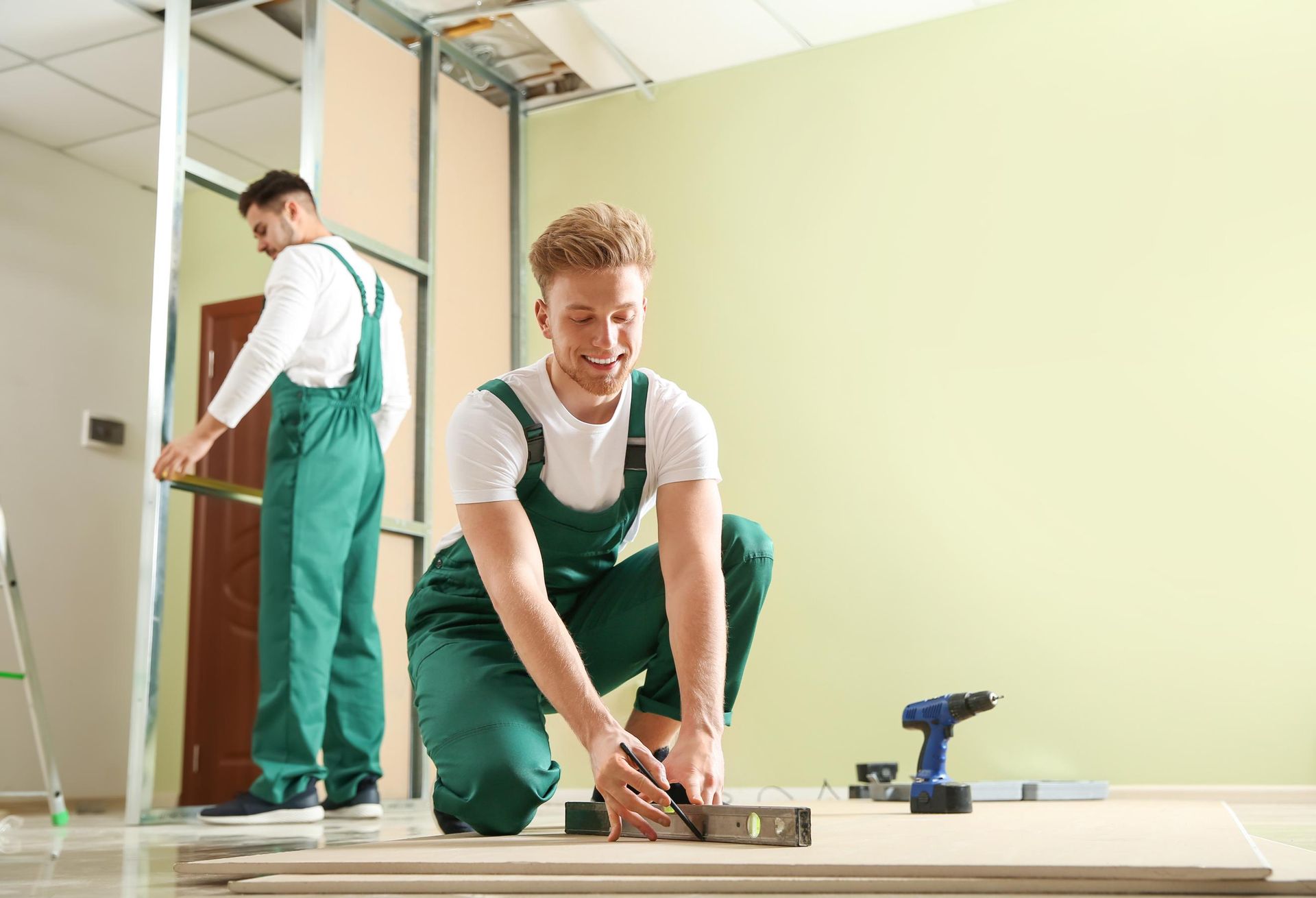 Two construction workers in green overalls; one kneels, measuring wood, while the other measures a wall frame.