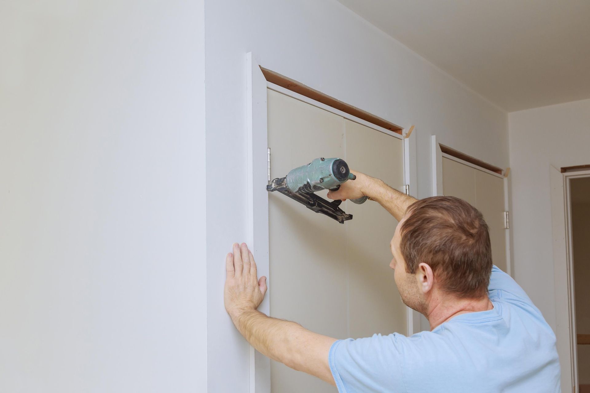 Man using a nail gun to install trim around a doorway in a white room.