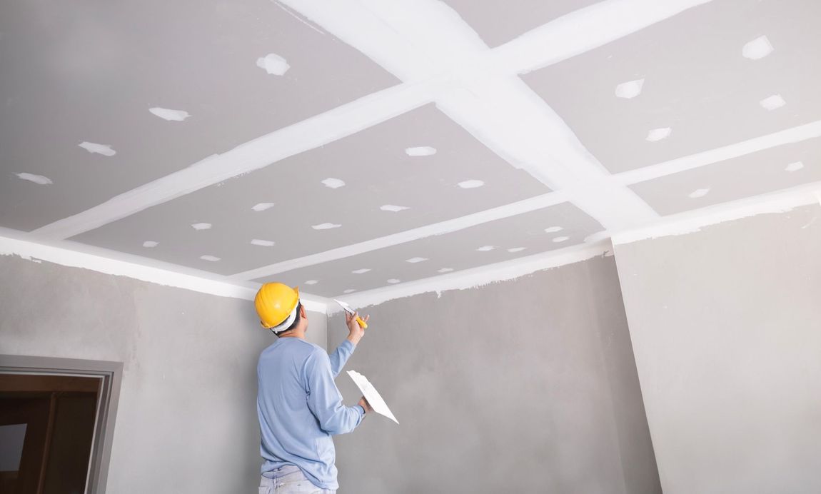 Construction worker in a yellow hard hat taping a drywall ceiling with white compound, inside a room.