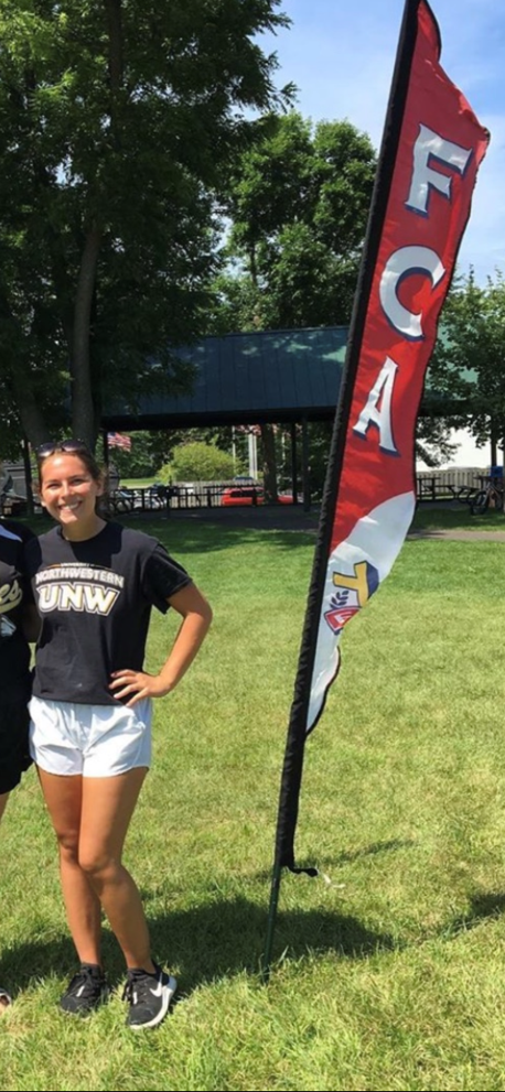 College-age female athlete standing next to FCA flag in grassy park with pavilion in the background.