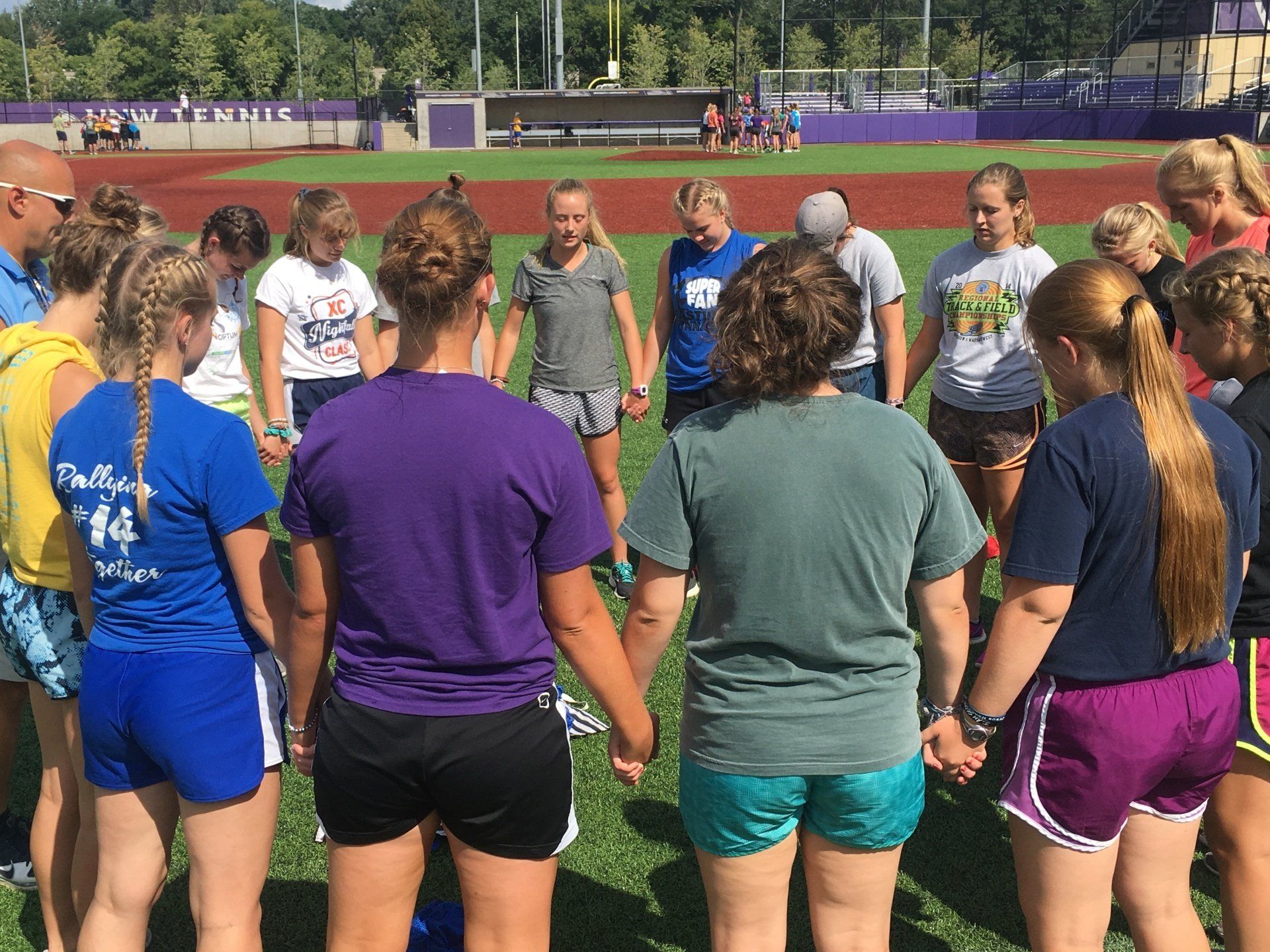 Female athletes standing in circle on baseball field holding hands in prayer.