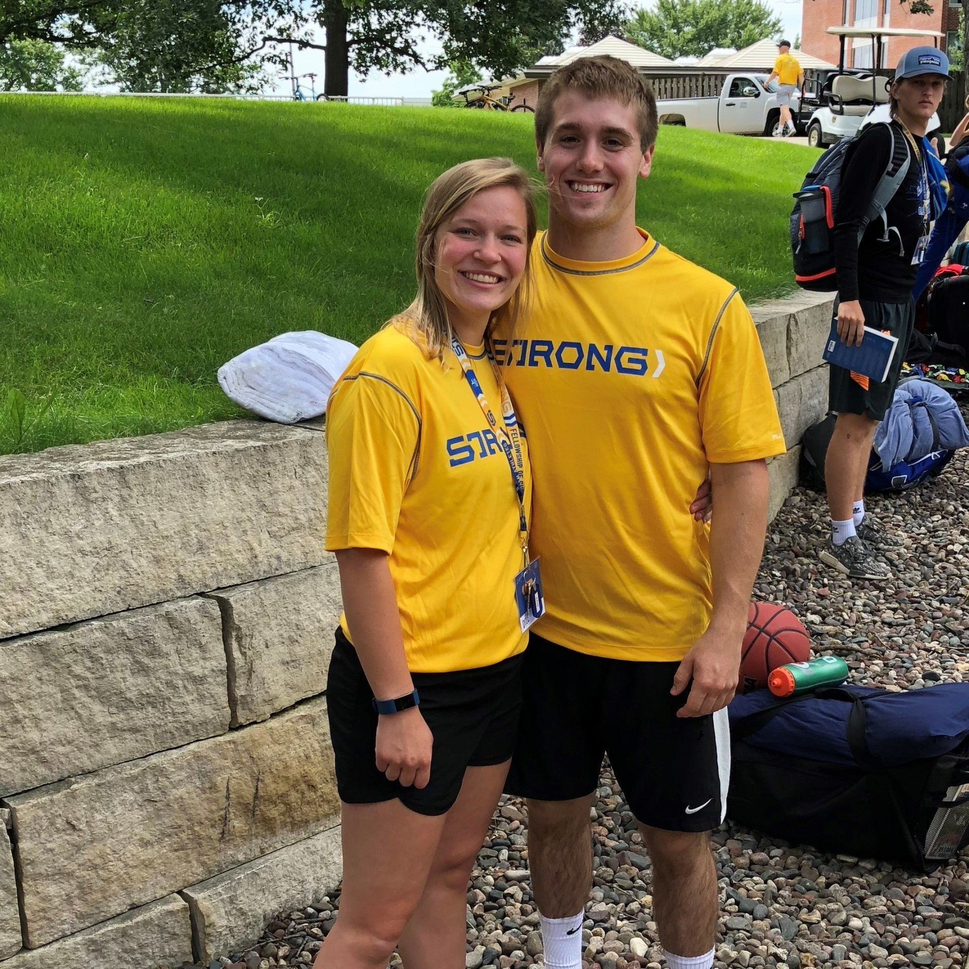 Two college age students wearing yellow FCA 'STRONG' t-shirts, standing outside with athletic equipment around them.