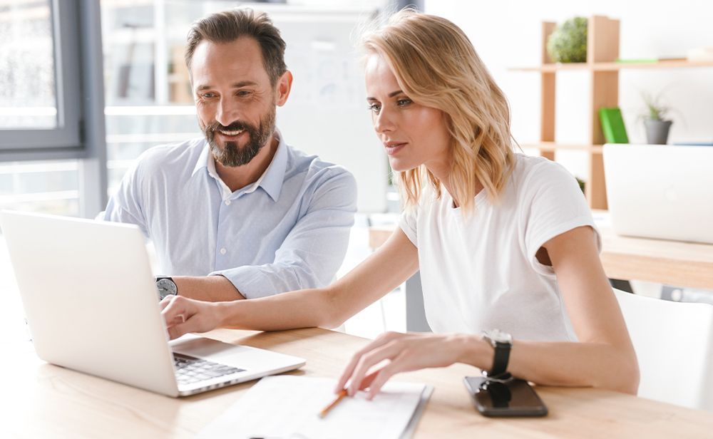 A man and a woman are sitting at a table looking at a laptop computer.