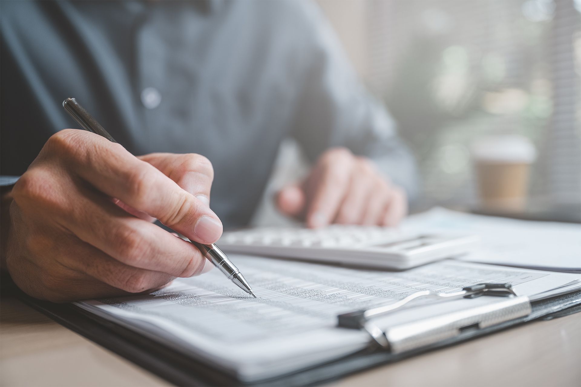 Person writing with a pen on paperwork with calculator, office setting.