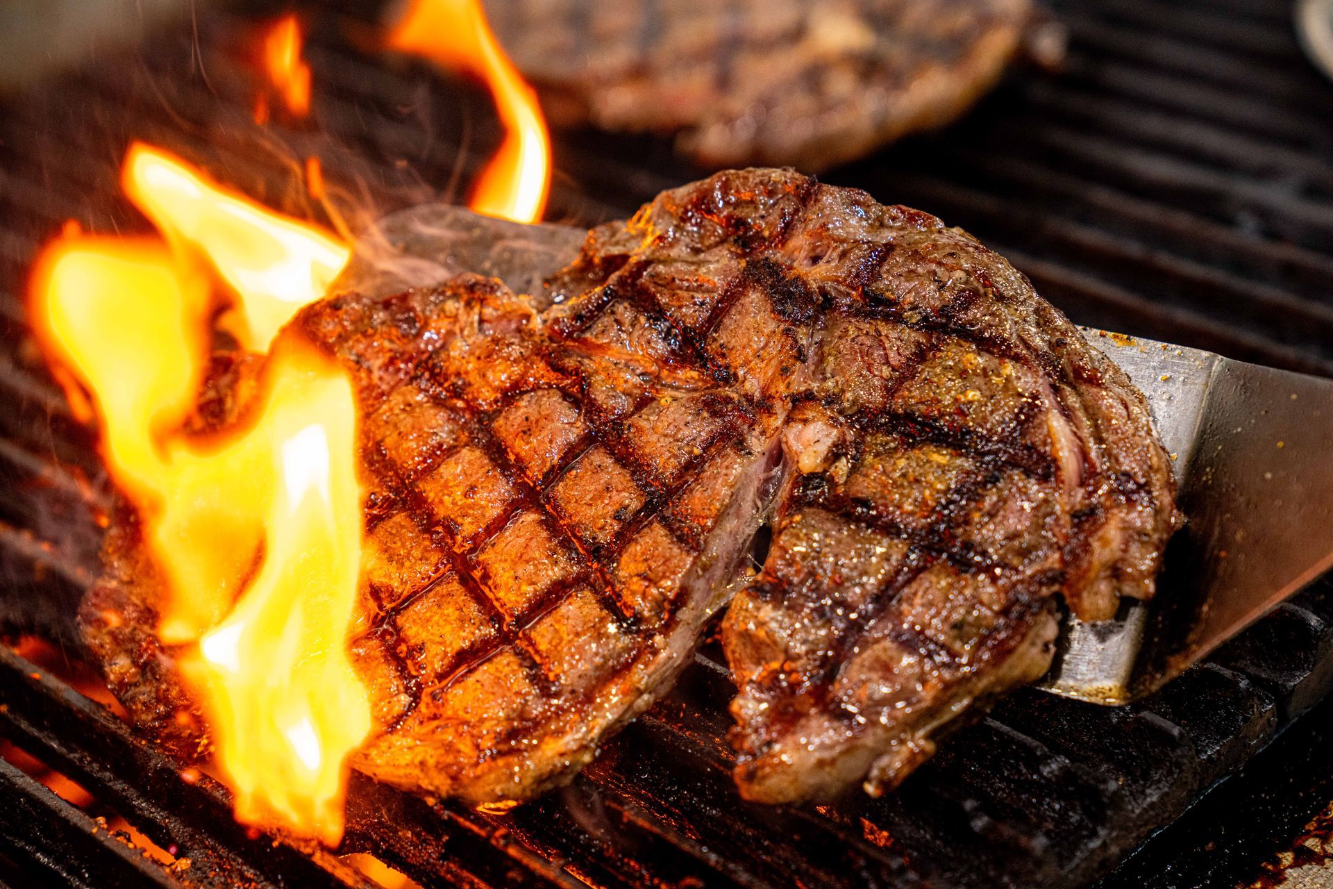 Two steaks are being cooked on a grill with flames behind them.