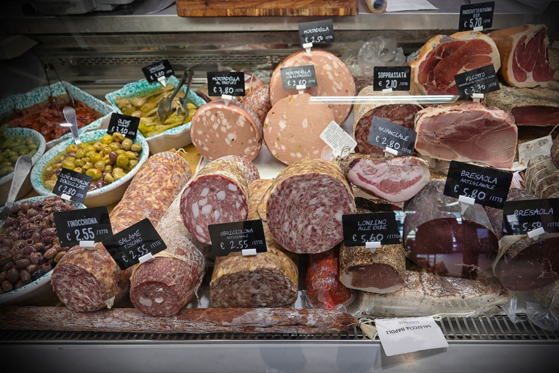 Banco gastronomia con esposizione di carni e olive di vario tipo, con cartellini dei prezzi.