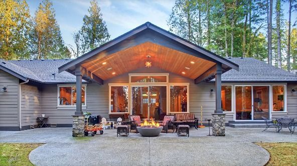 Back view of a house with a large wooden covered patio, a stone fire pit, outdoor seating, and surrounding trees.