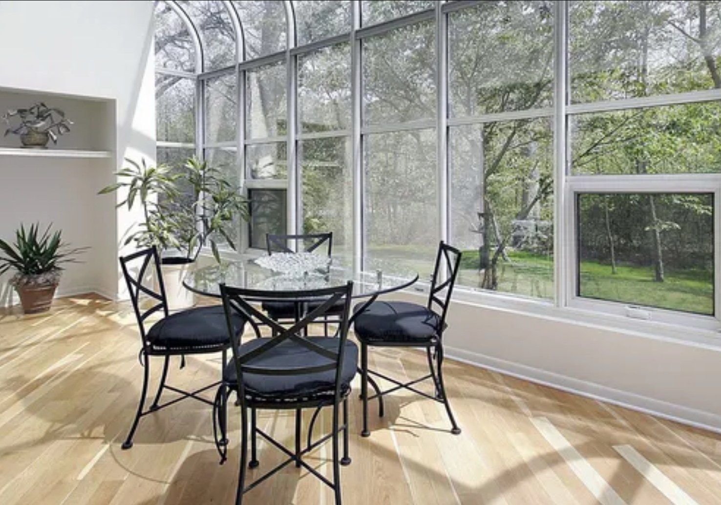 A bright sunroom with a glass dining table, four black metal chairs, light wood floors, and floor-to-ceiling windows.