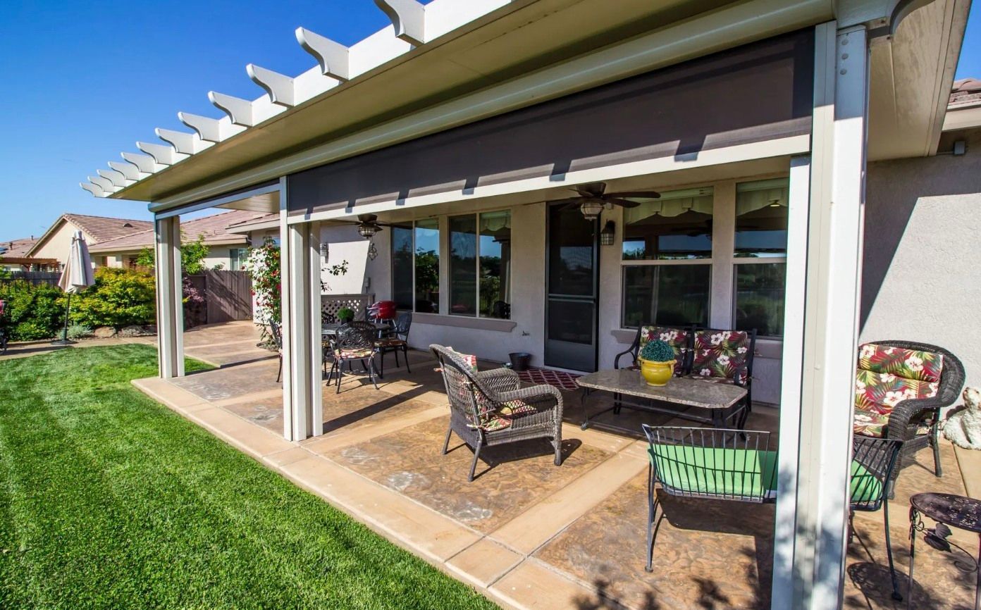 A shaded outdoor patio with wicker furniture, a table, and a green lawn under a sunny sky.