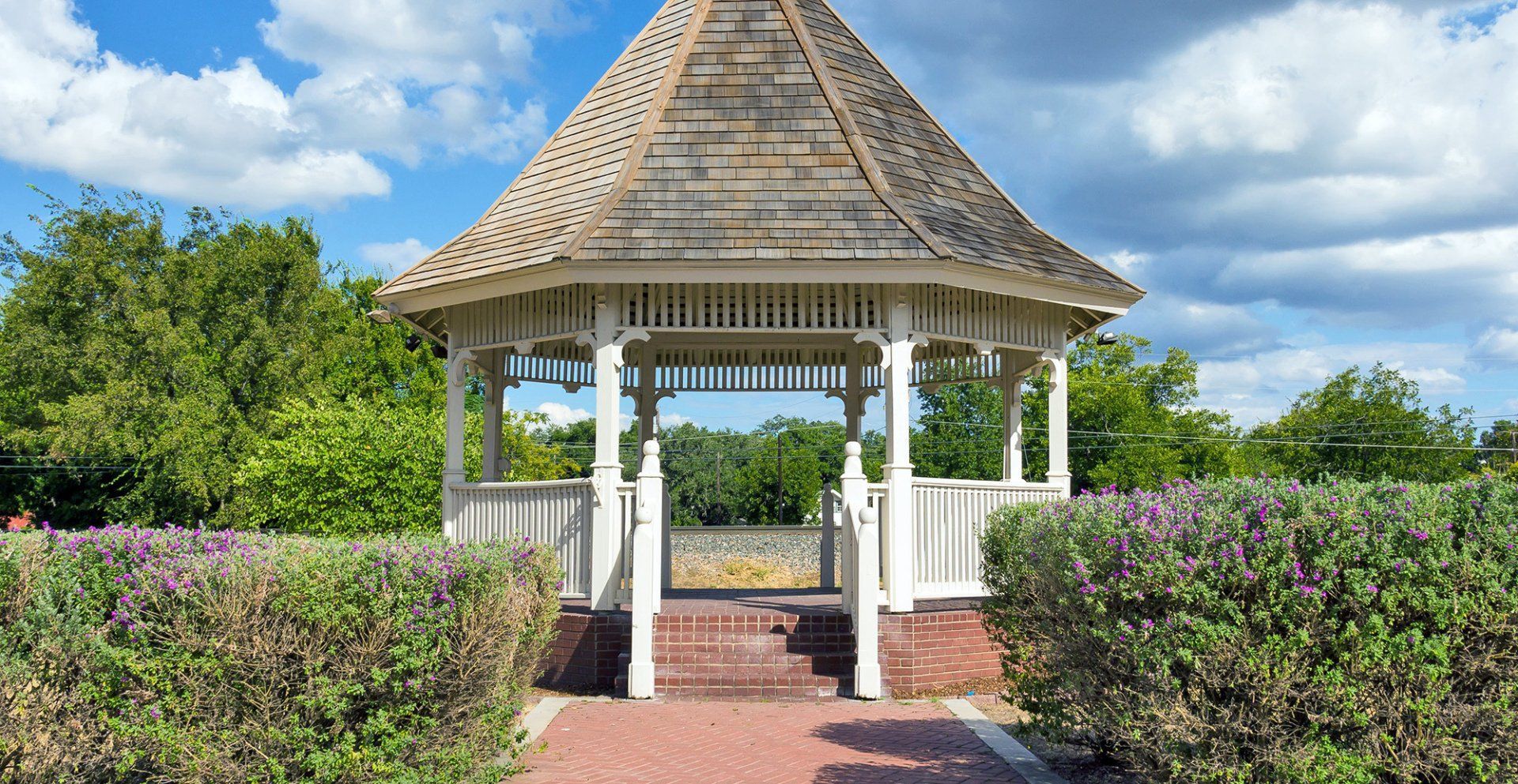 A white wooden gazebo with a shingled roof stands at the end of a brick path, surrounded by bushes under a blue sky.