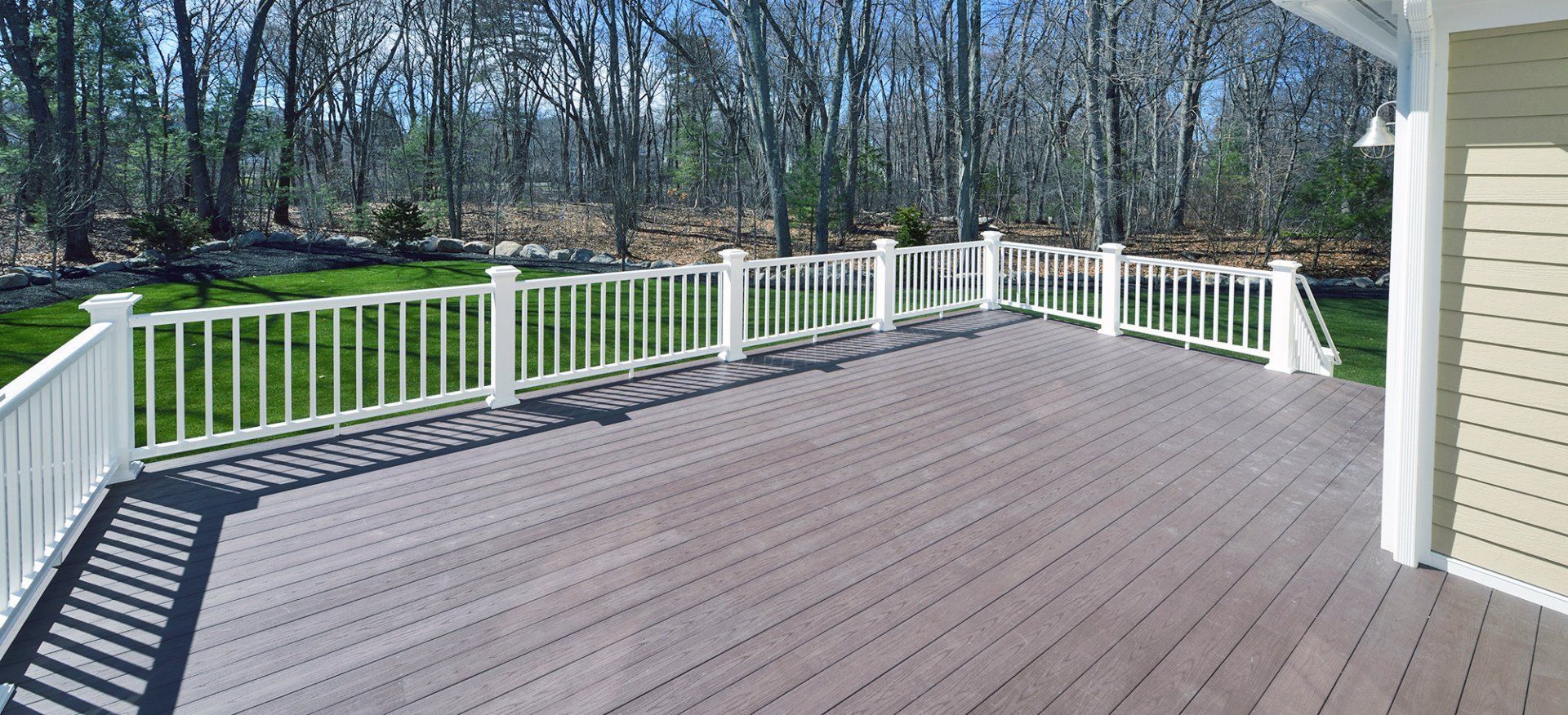 A high-angle view of a grey composite deck with white railings, overlooking a grassy yard with trees in the background.