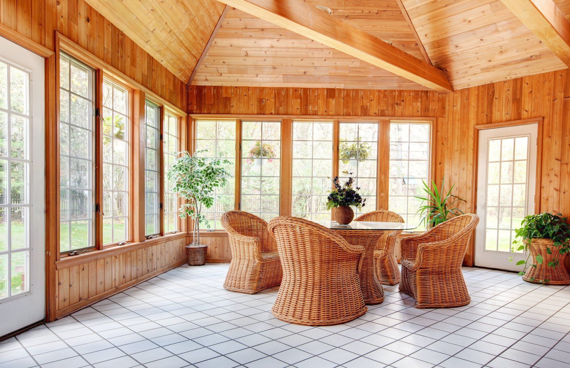 A sunroom with natural wood walls, tiled floor, a wicker dining set, and large windows overlooking greenery.
