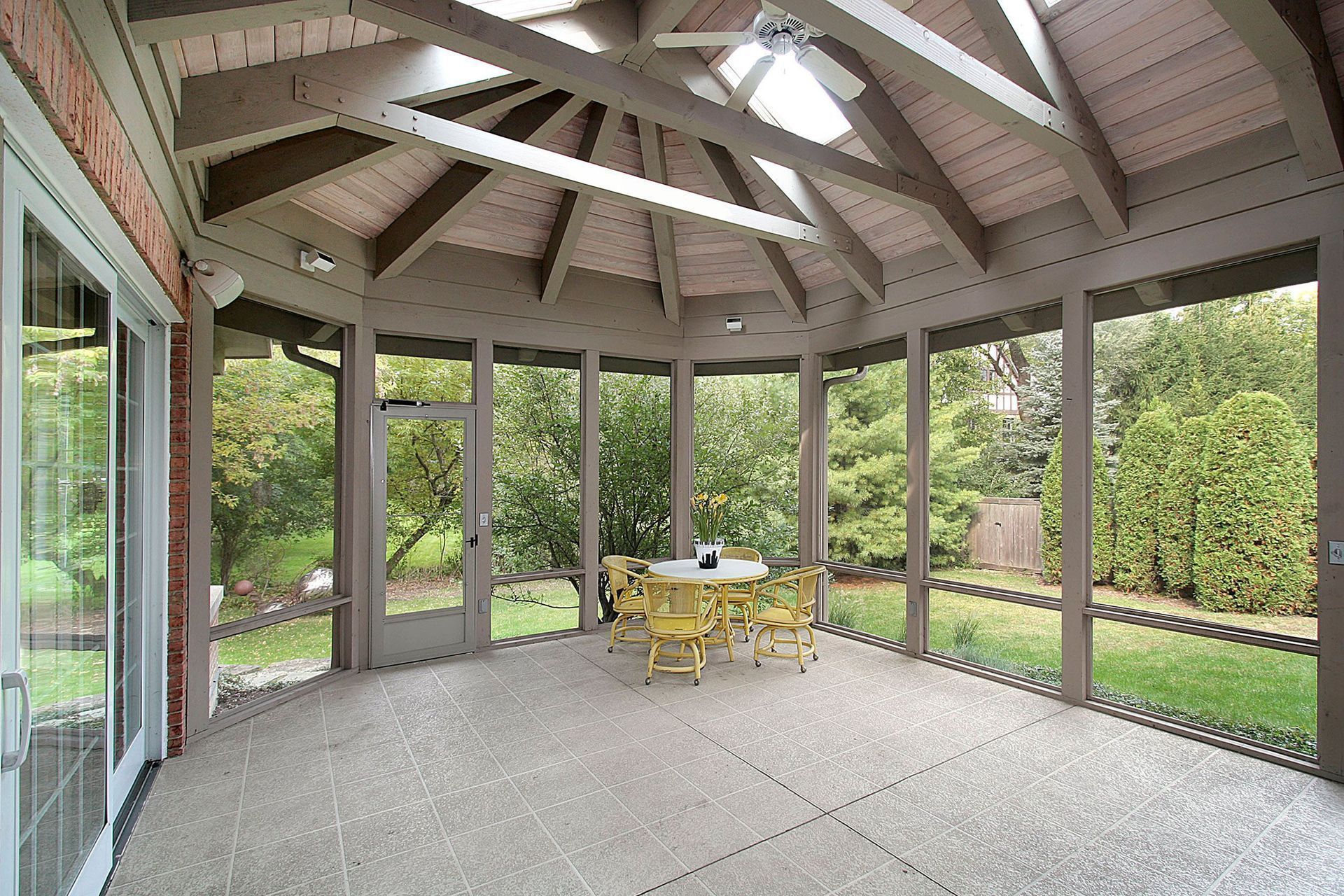A sunroom with a vaulted wooden ceiling, screened walls looking out to a garden, and a small table with yellow chairs.