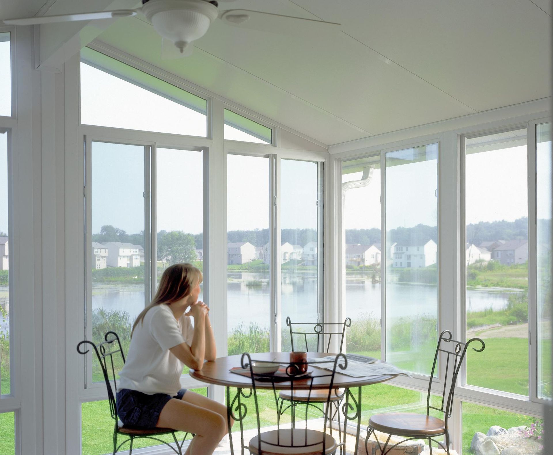 A person sits at a table in a sunroom, looking out large windows at a pond and residential neighborhood.