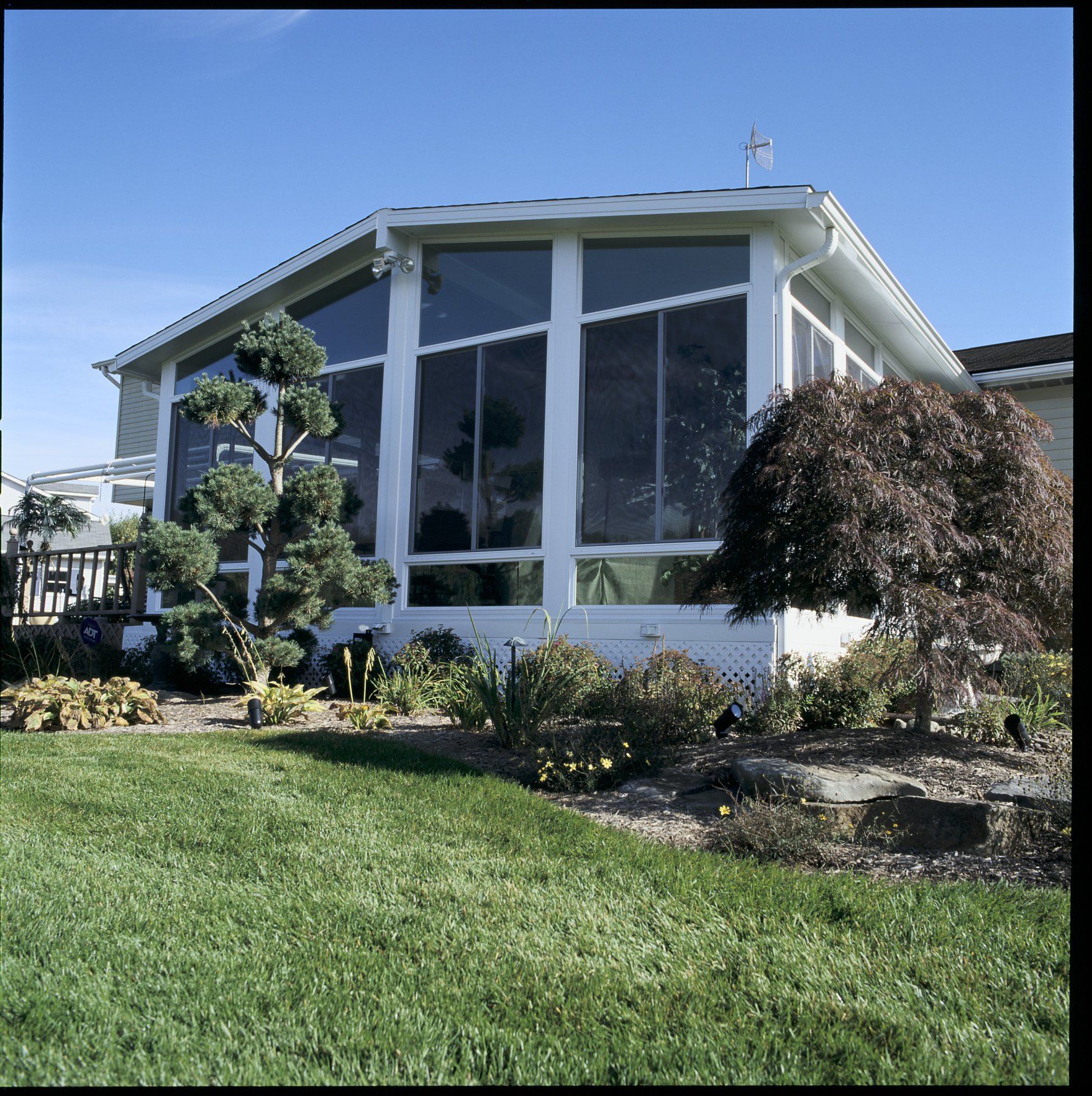 A white sunroom addition with large glass windows and a peaked roof, set in a yard with trees and green grass.