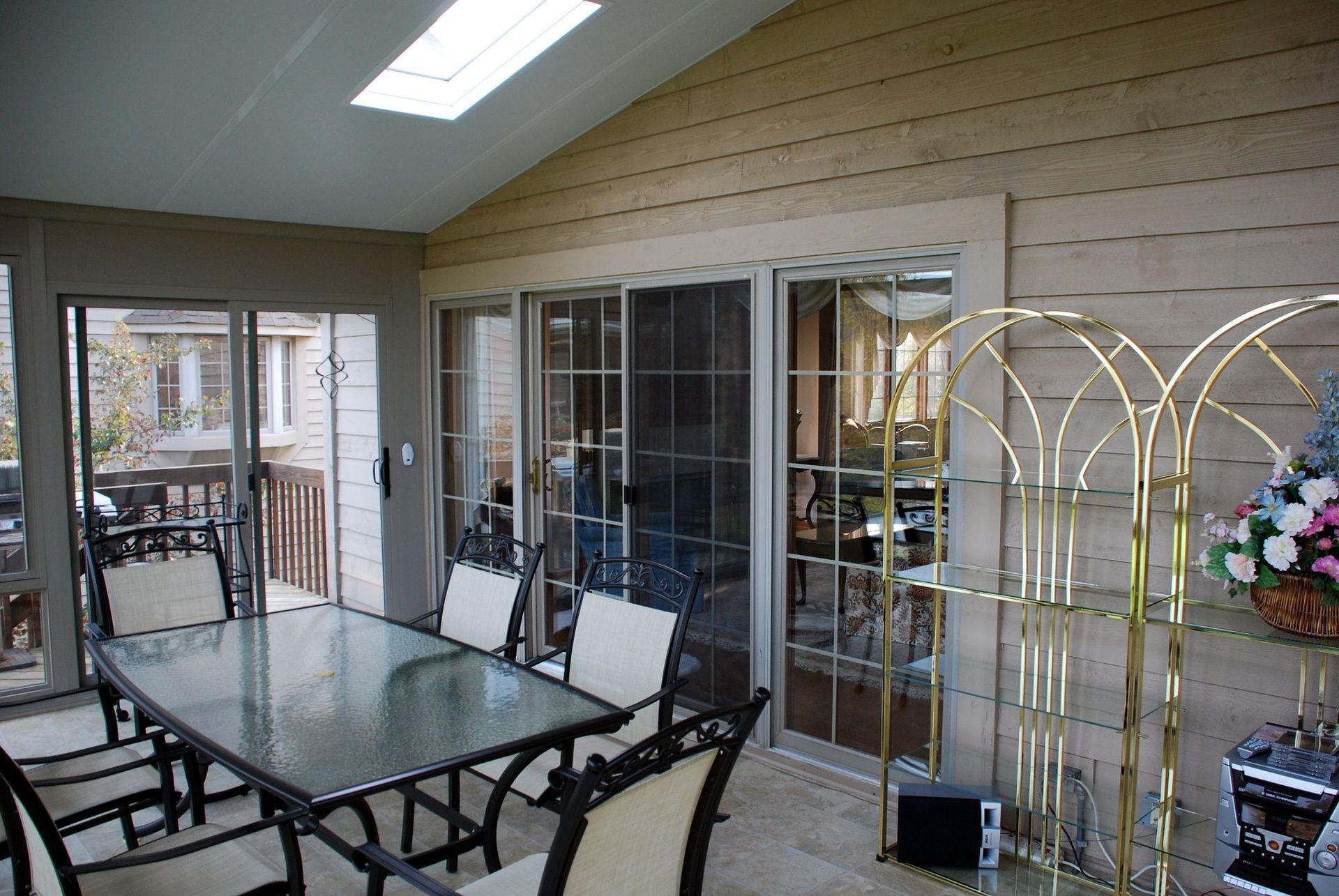 A sunroom with a glass dining table, four chairs, a tall gold-toned shelving unit, and large sliding glass doors.