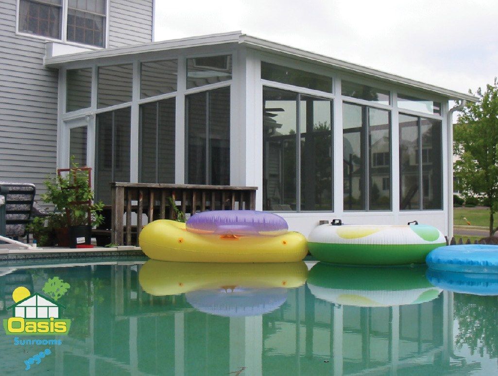 A white sunroom addition with glass walls stands next to a pool with three colorful floating rings on the water.
