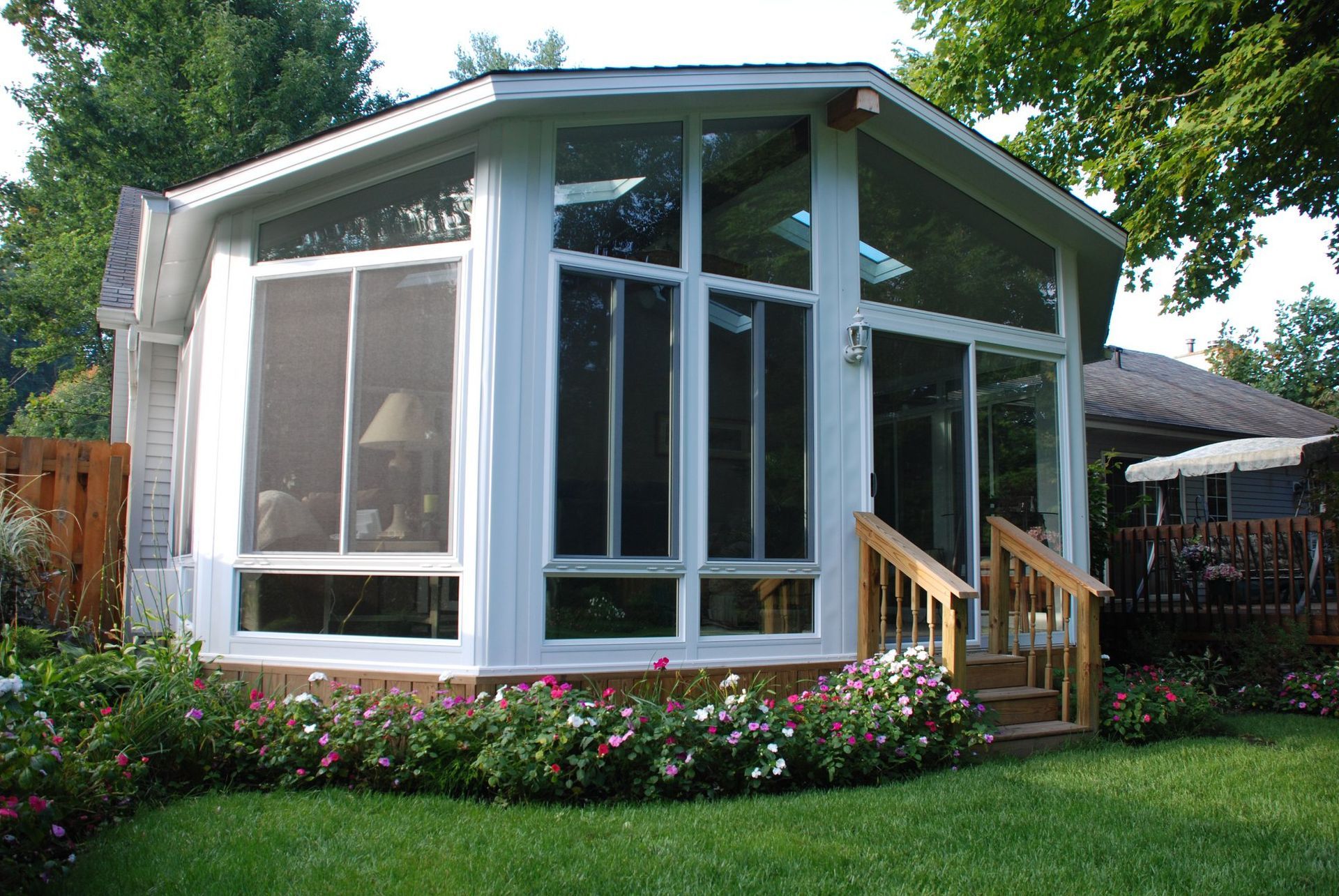 A white, multi-windowed sunroom addition sits in a lush green backyard, bordered by a bed of colorful flowers.