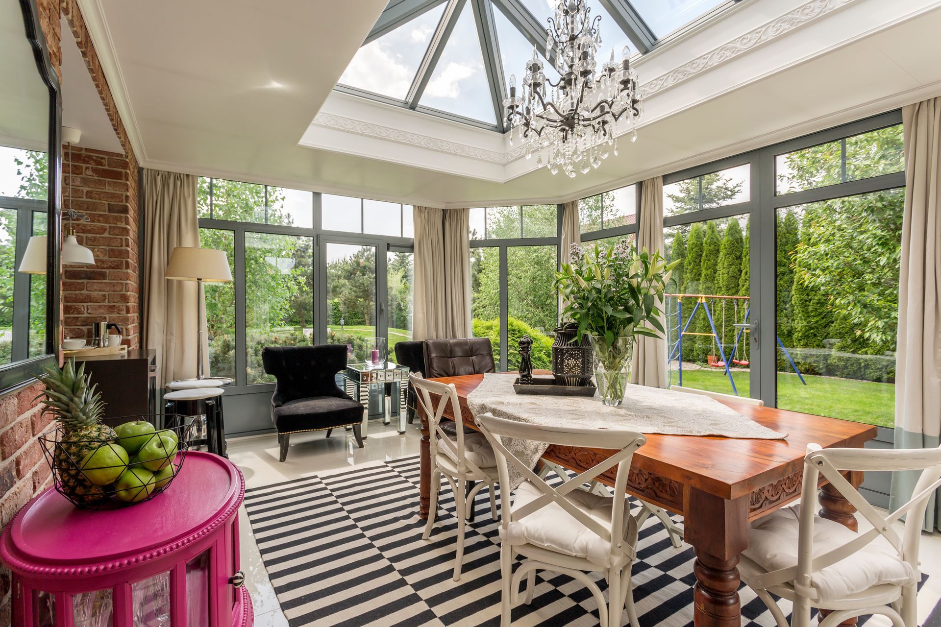 A sunroom featuring a dining table with white chairs, a black-and-white striped rug, and a vibrant pink side table.