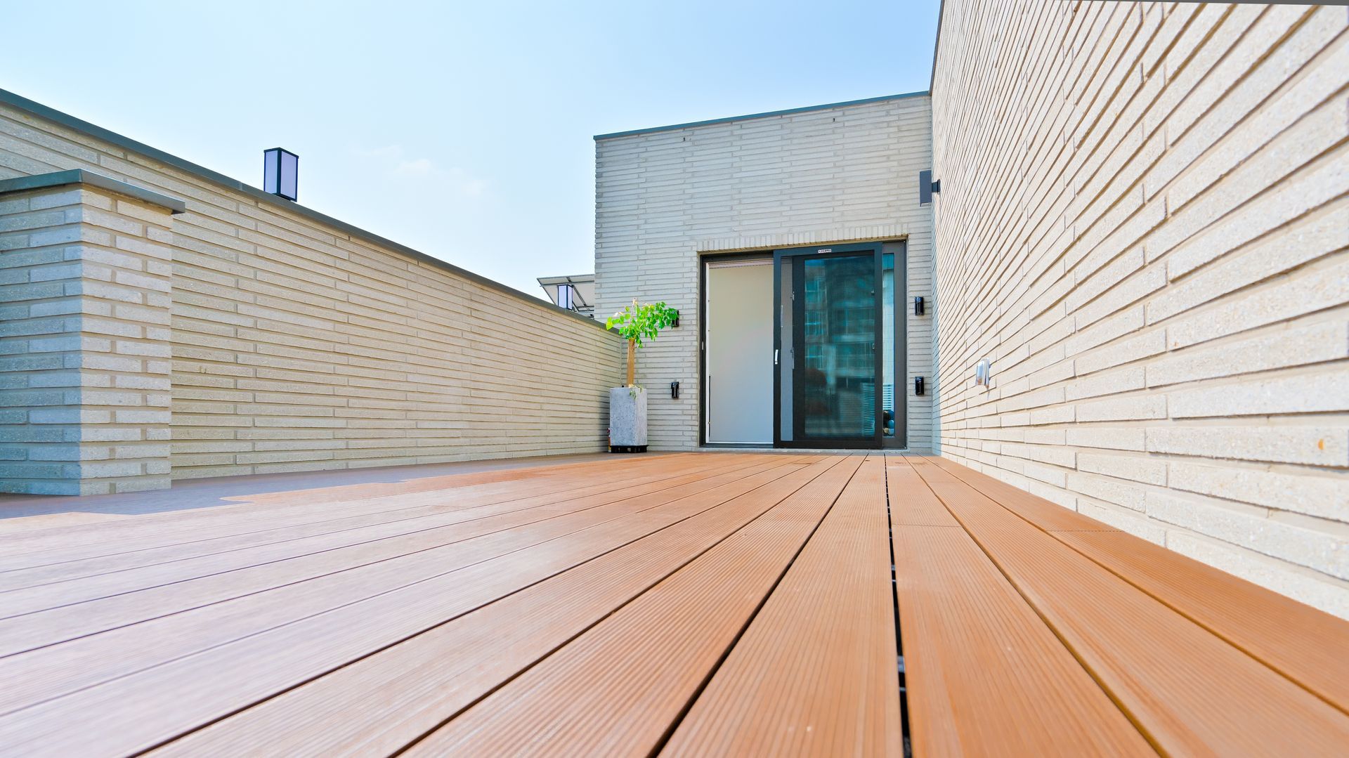 A wide-angle view of a wooden rooftop deck leading to a glass sliding door between two textured, light-colored walls.