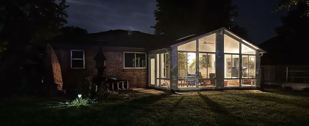 A bright sunroom with a glass dining table, four black metal chairs, light wood floors, and floor-to-ceiling windows.