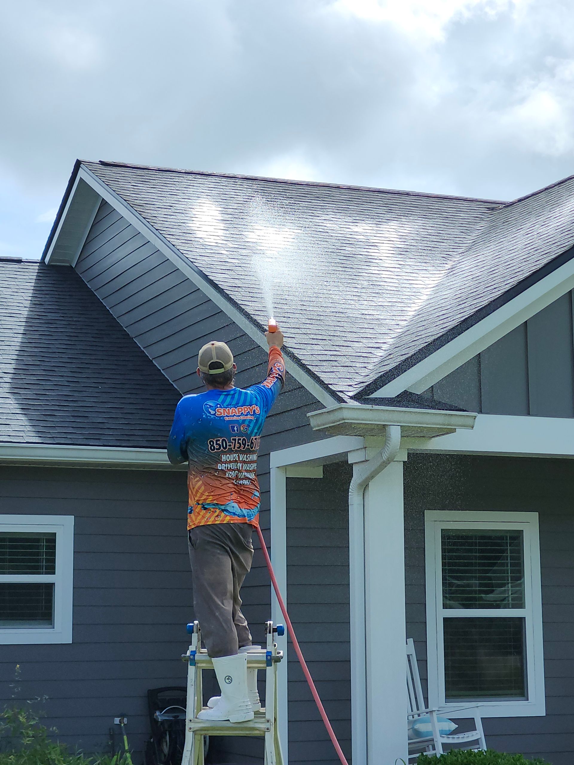 Man power washing a gray shingled roof from a ladder on a cloudy day.