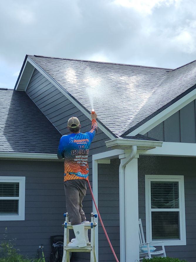 Man power washing a gray shingled roof from a ladder on a cloudy day.
