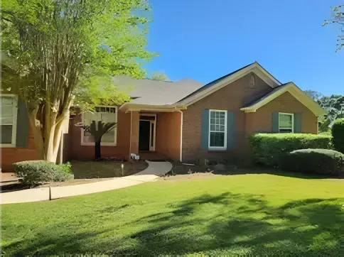 Brick house with green shutters, a curved walkway, and a well-manicured lawn under a bright blue sky.