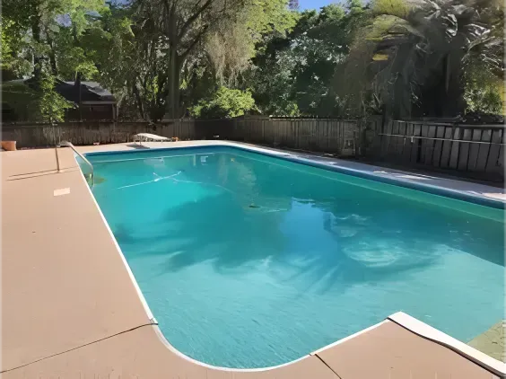 Pool with blue water surrounded by a concrete deck. Fenced backyard, trees, and blue sky in the background.
