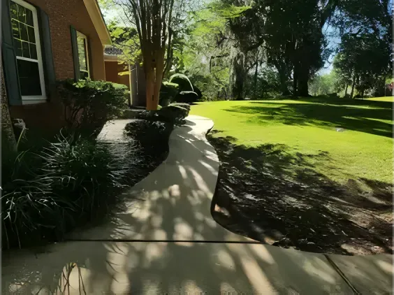 A paved walkway curves through a green lawn towards a house with trees and blue shutters on a sunny day.