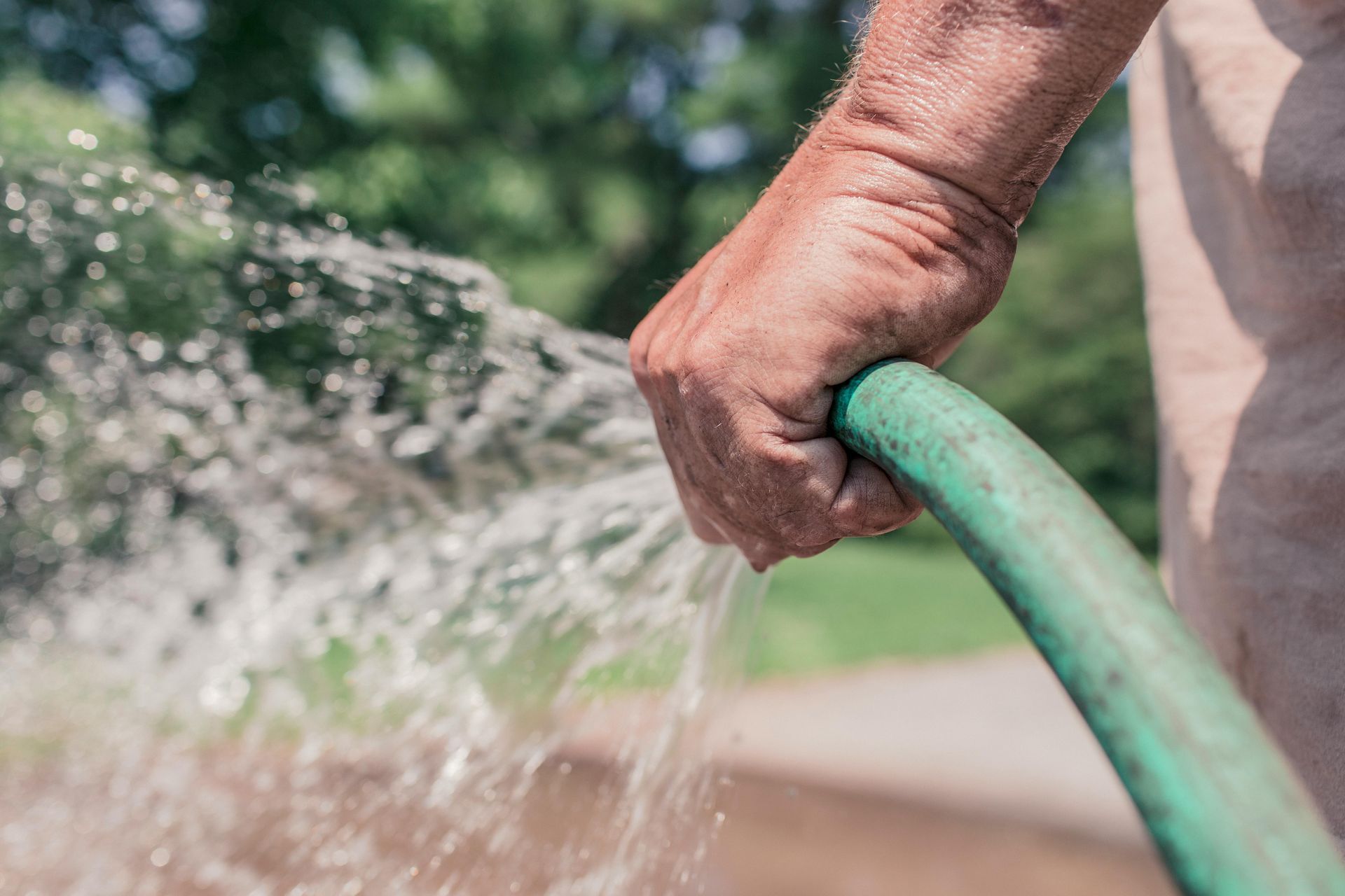 Man's hand holding a green hose, spraying water outdoors on a sunny day.