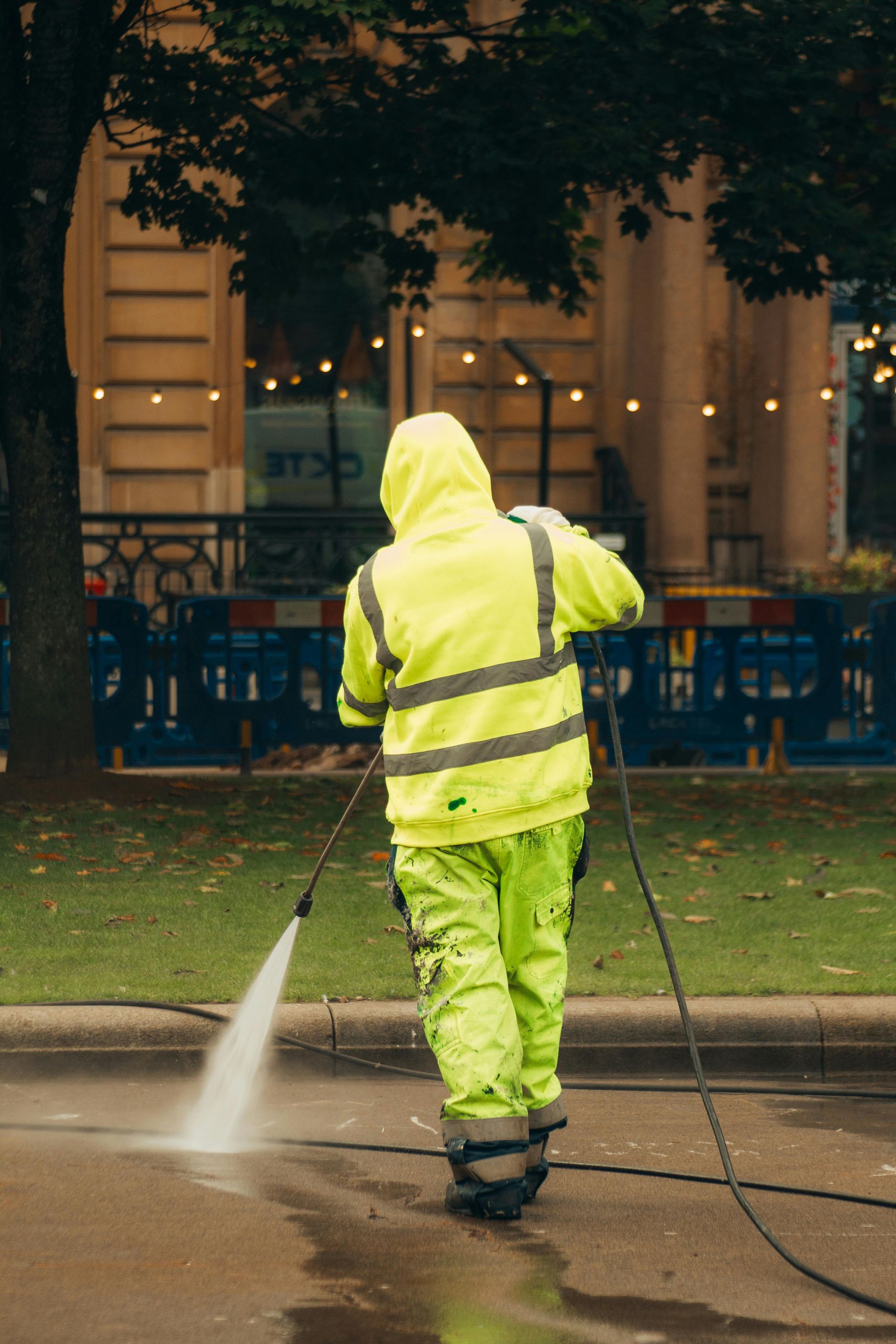 Person in neon yellow rain gear power washing a street, near a building.