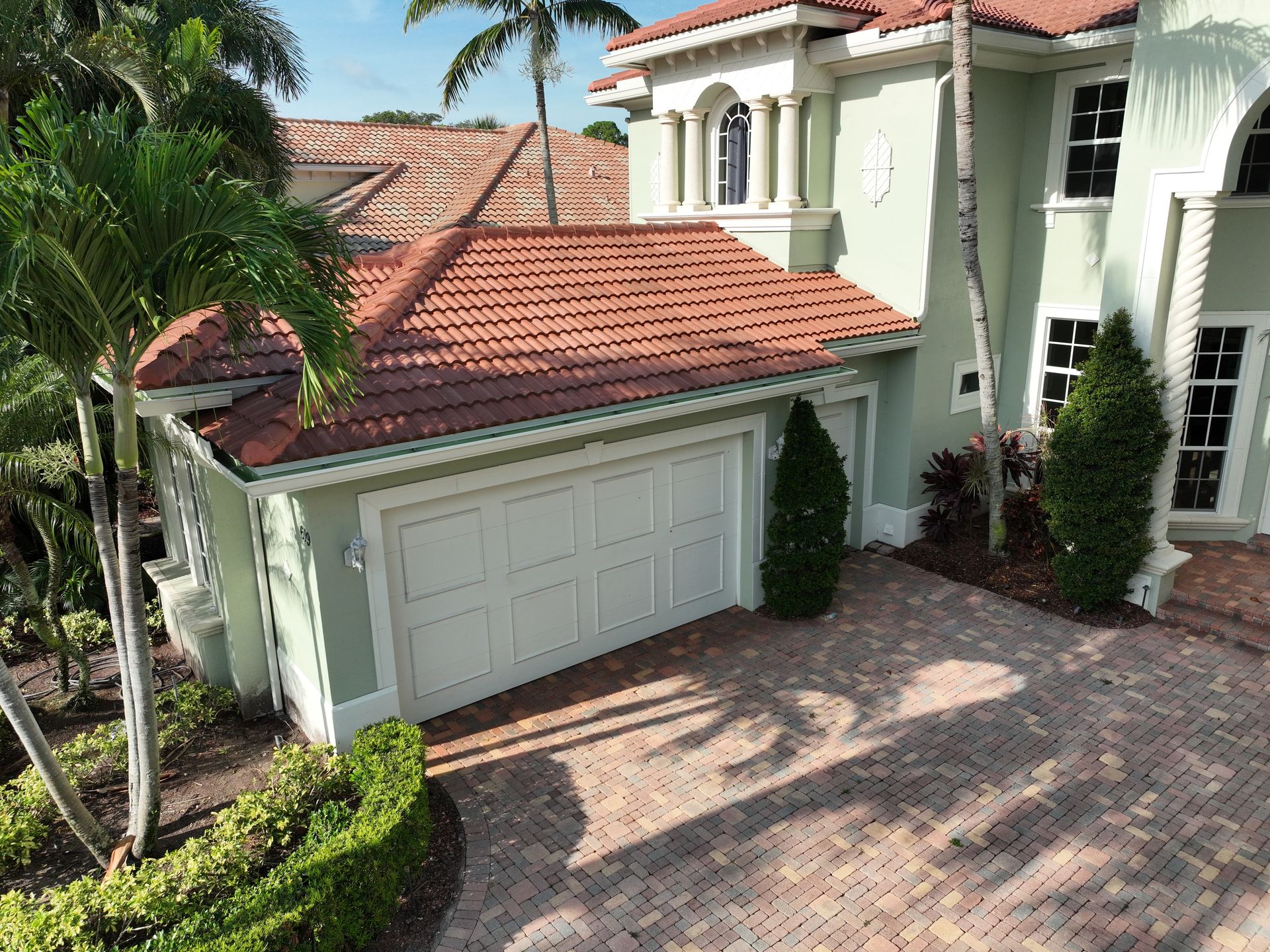 A light green house with a red tile roof and a white garage door sits on a brick driveway.