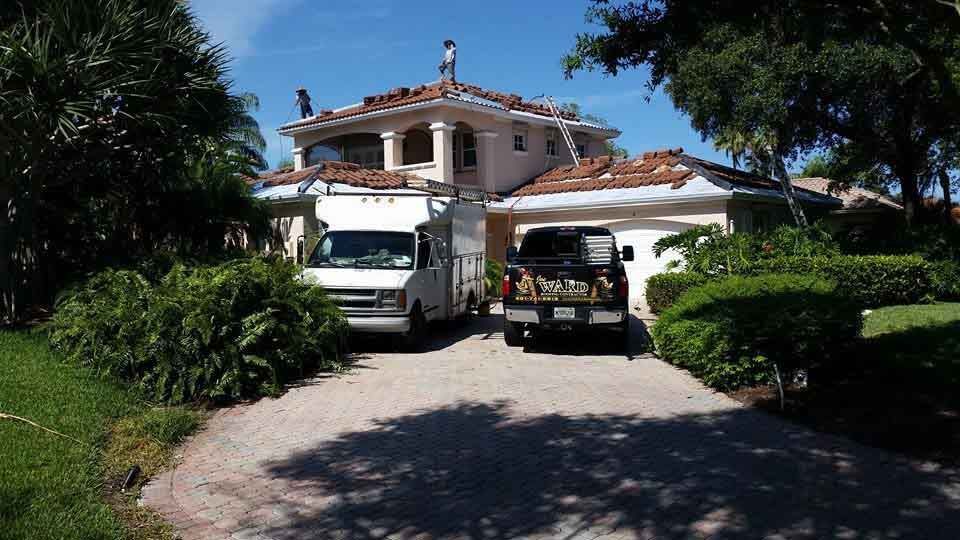 Roofers working on a two-story beige house with a clay tile roof; vehicles in the driveway.