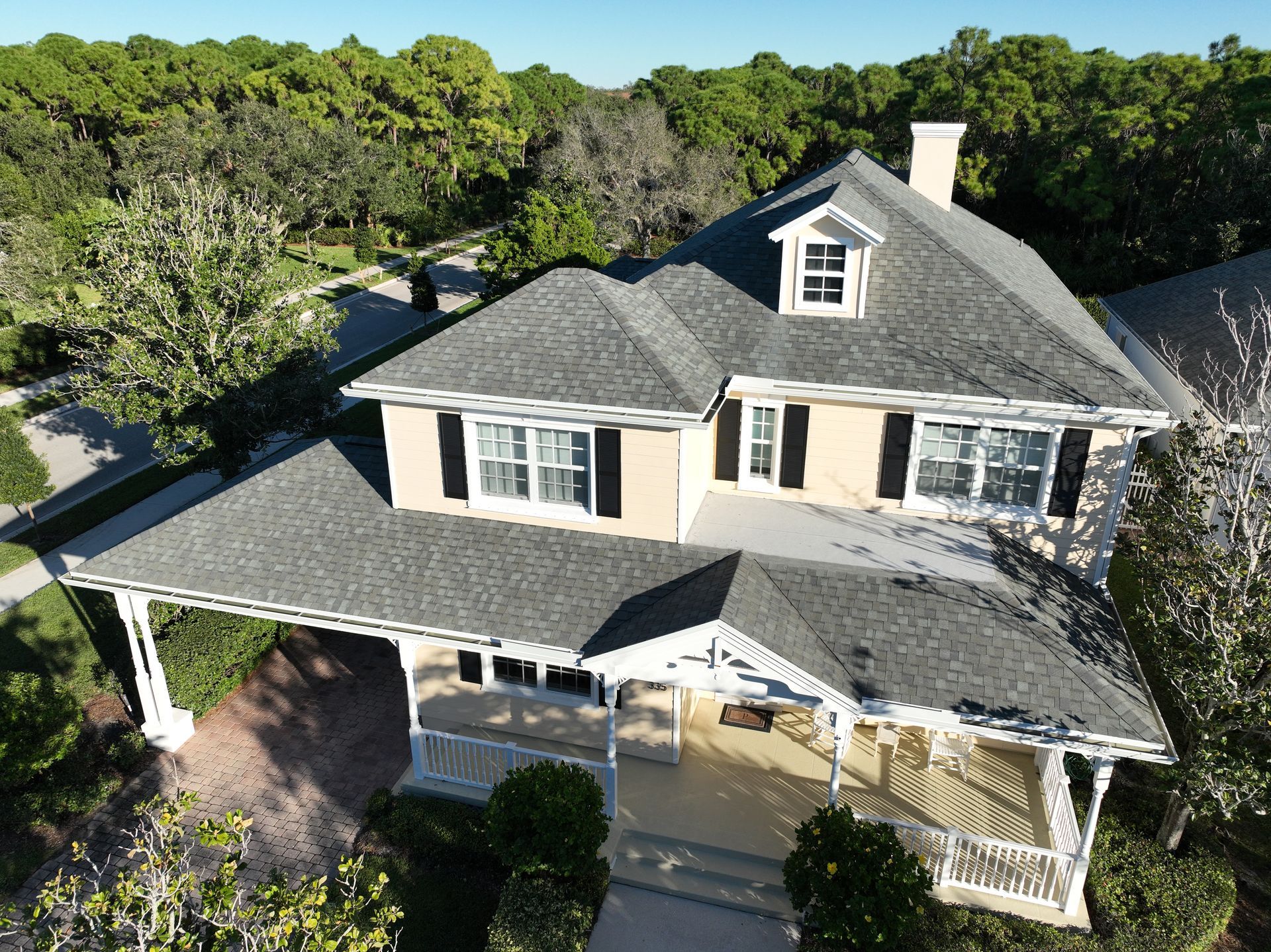 An aerial view of a light-colored house with a dark gray roof, surrounded by trees and greenery.
