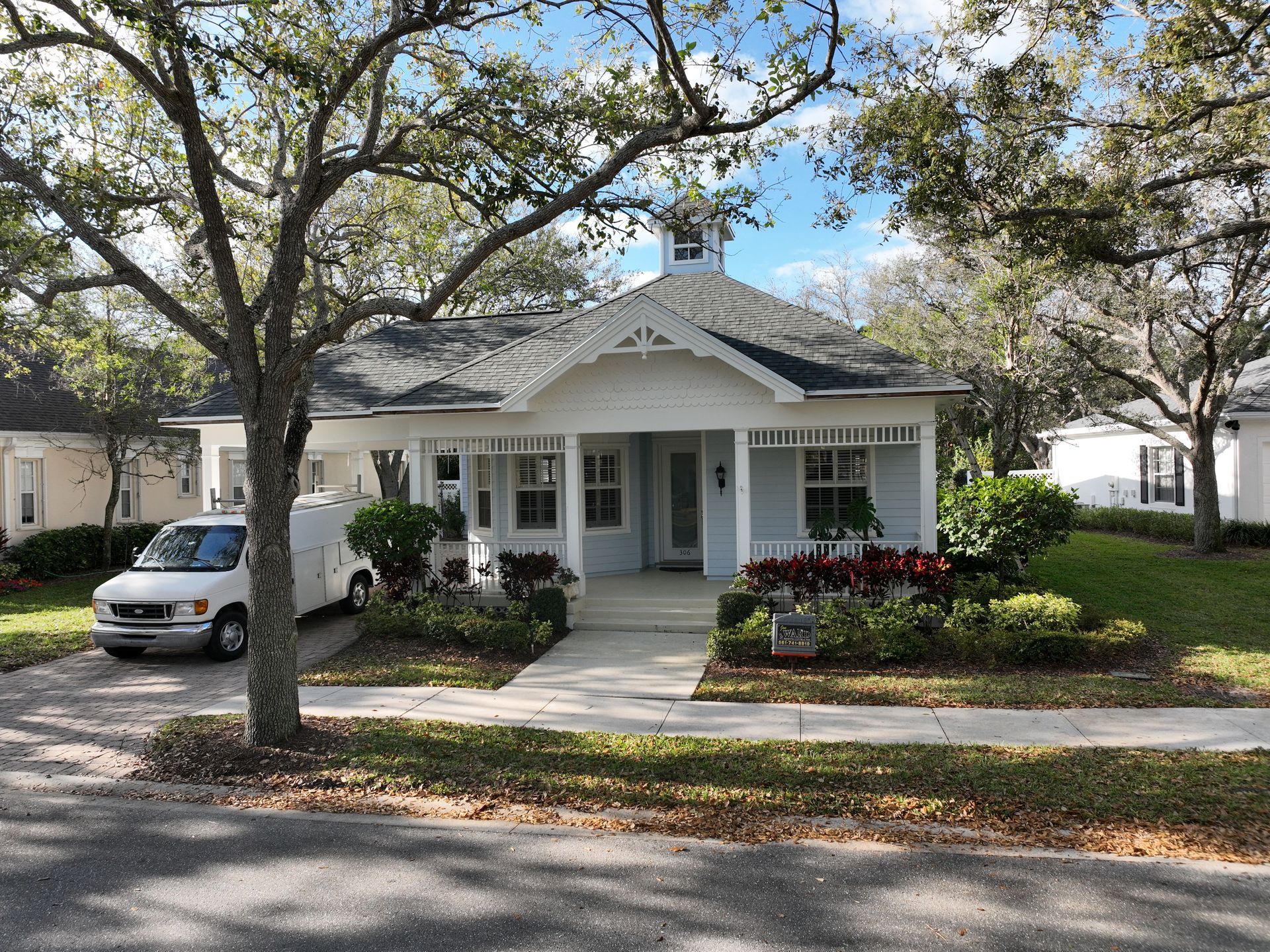 Blue and white cottage with porch, flowerbeds, parked van, and trees on a sunny day.