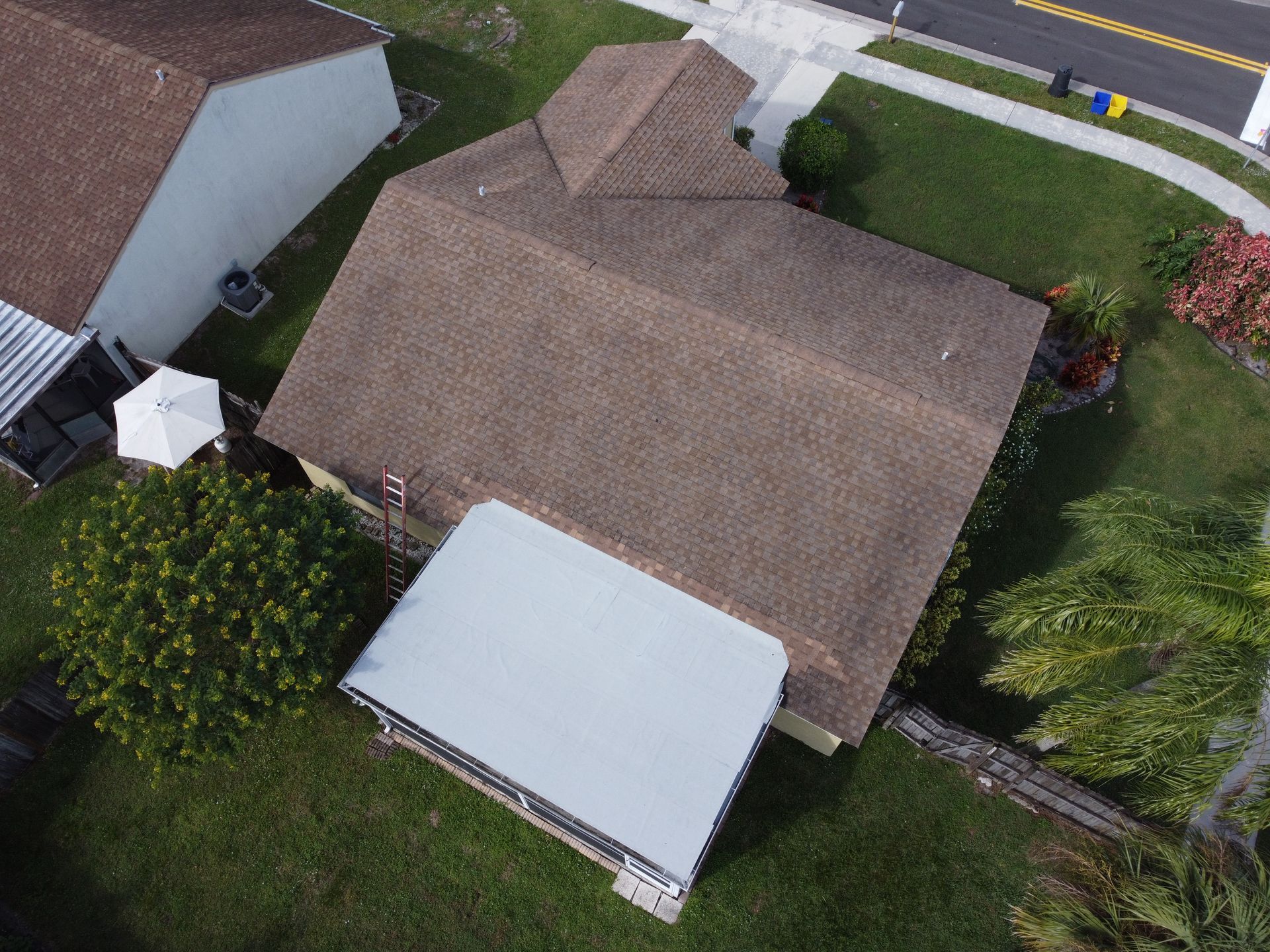 Aerial view of a house with brown roof, white shed, and green lawn in a neighborhood.