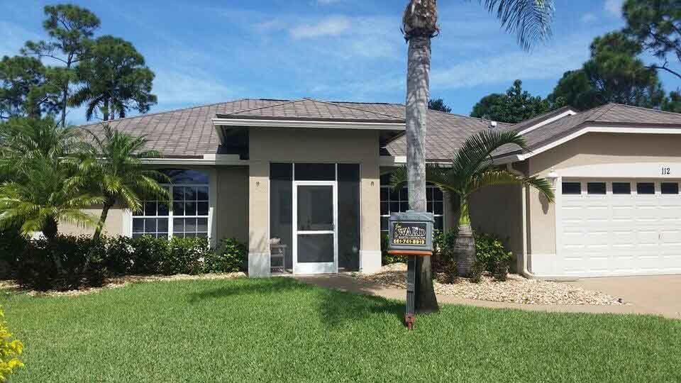 Beige house with front door under a porch, surrounded by green lawn and palm trees.