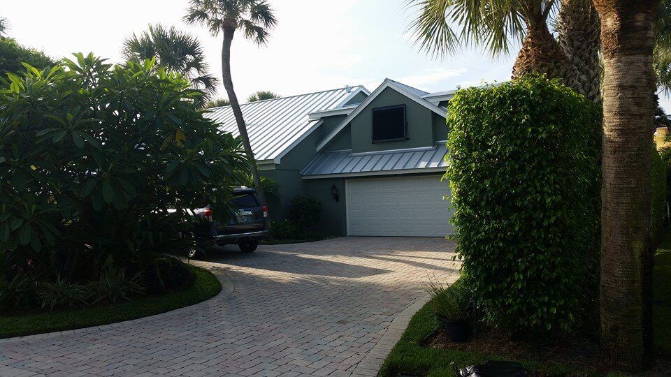 A house with green siding and a silver roof, white garage door, and a brick driveway, surrounded by trees.