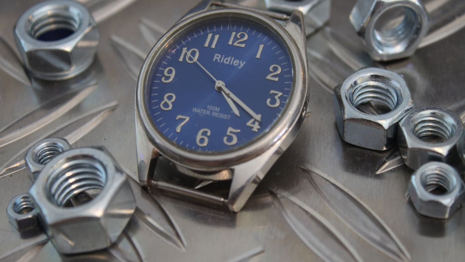 Reloj de pulsera con esfera azul rodeado de tuercas metálicas sobre una superficie plateada.