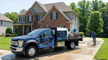 A blue work truck washing a driveway in front of a two-story house; a worker sprays the concrete.