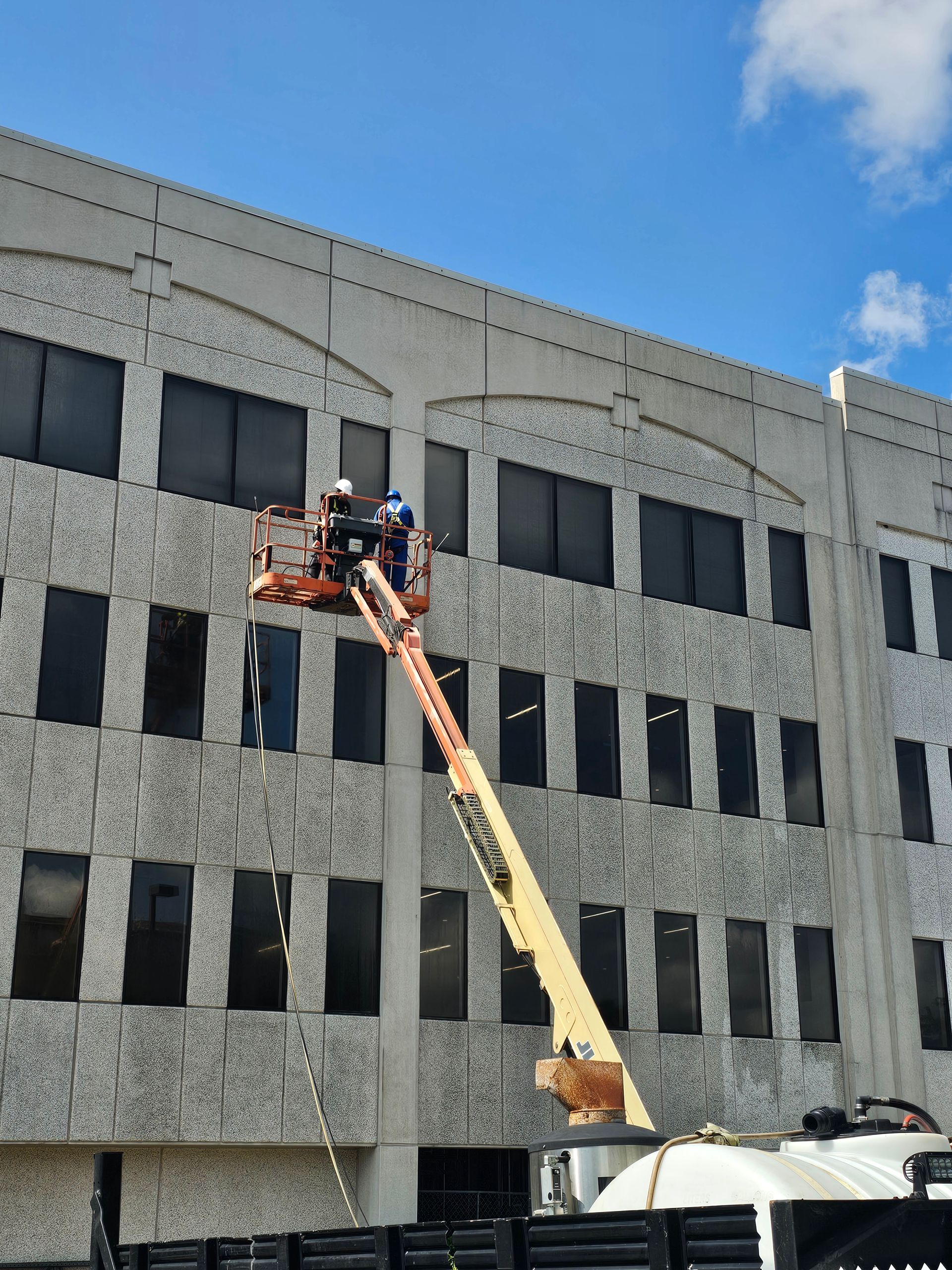 A blue work truck washing a driveway in front of a two-story house; a worker sprays the concrete.