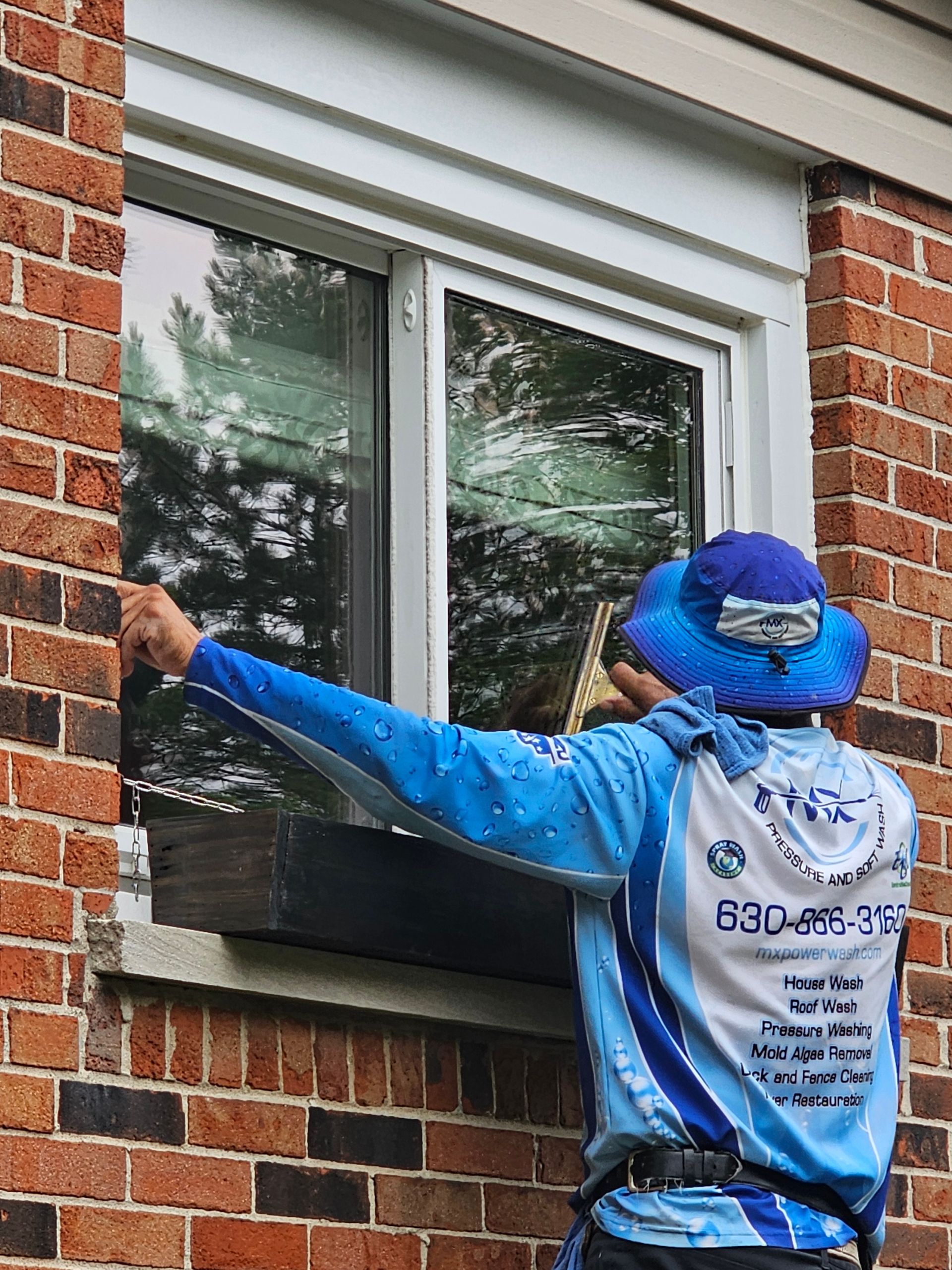 A blue work truck washing a driveway in front of a two-story house; a worker sprays the concrete.