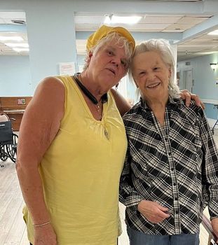Two women smiling and embracing in a light-filled room. One wears yellow, the other a plaid shirt.